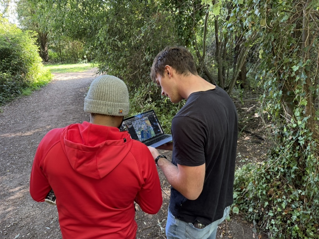 [Photo of researchers consulting a laptop on a country road surrounded by green overgrowth.]